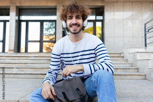Happy young university student sitting on stairs in campus wearing headphones looking at camera.