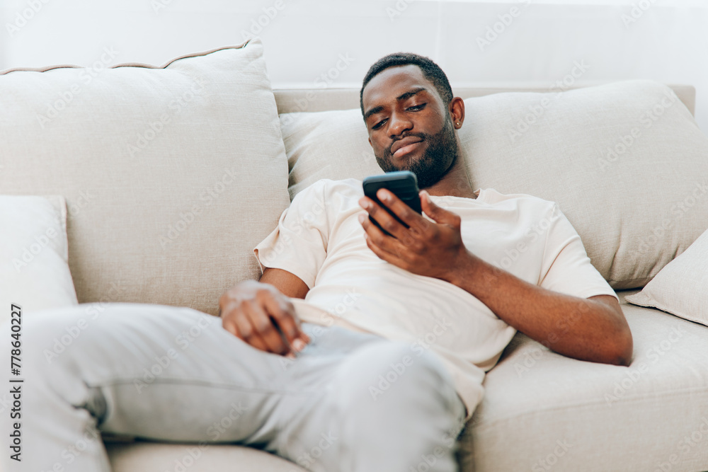Happy African American man sitting on a black sofa, holding a mobile phone, typing a message He is enjoying his leisure time at home, connected to the internet, and embracing modern technology The Happy African American man sitting on a black sofa, holding a mobile phone, typing a message He is enjoying his leisure time at home, connected to the internet, and embracing modern technology The