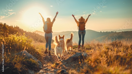 Two hikers hiking on mountain pathway with two dogs during sunset female friends hands in the air happy celebrate success reach destination photo