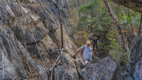 A man with a backpack and safety equipment climbs a cliff. With a gloved hand, he holds on to a gray rough steep slope. In the distance, from the back, another person among the branches of the trees. photo