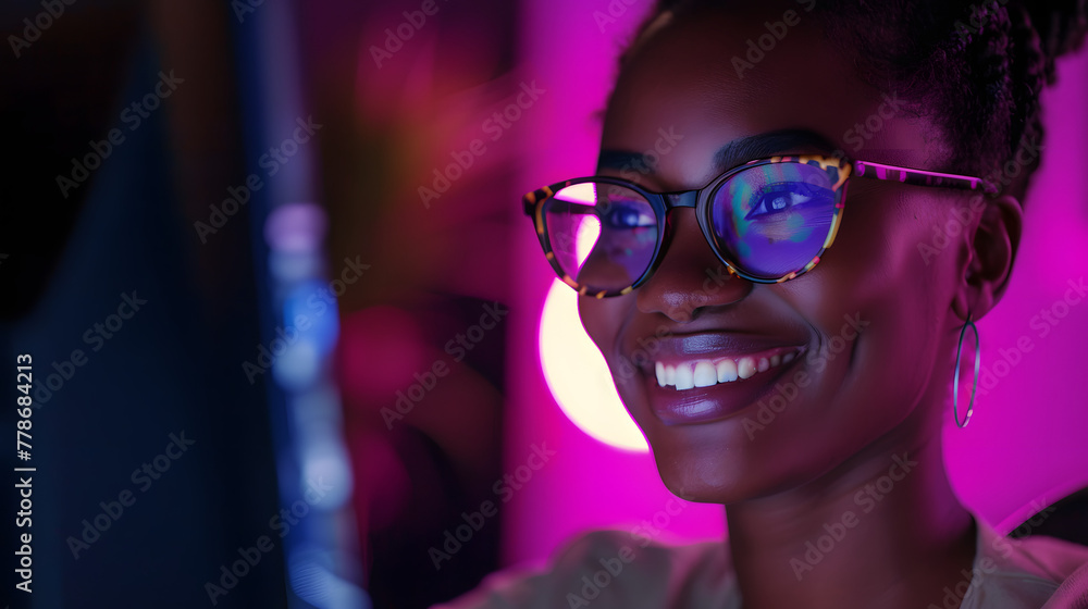A black woman wearing glasses is smiling while sitting in front of her computer screen