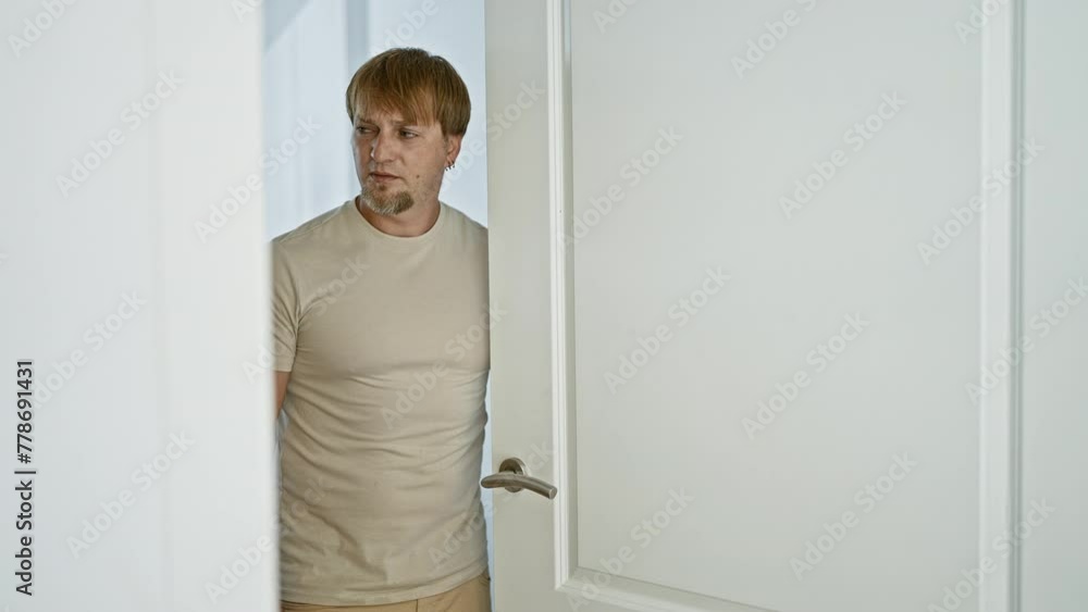 Bearded blond man in casual clothing inside a modern white interior room, looking pensive and alone. Bearded blond man in casual clothing inside a modern white interior room, looking pensive and alone.