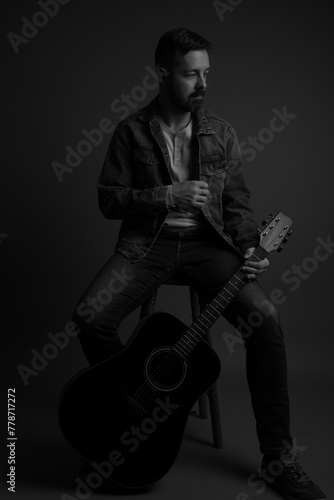 Black and white studio portrait of a handsome caucasian man sitting on a stool and holding an acoustic guitar. He is wearing running shoes, jeans, jean jacket and a white shirt underneath. 