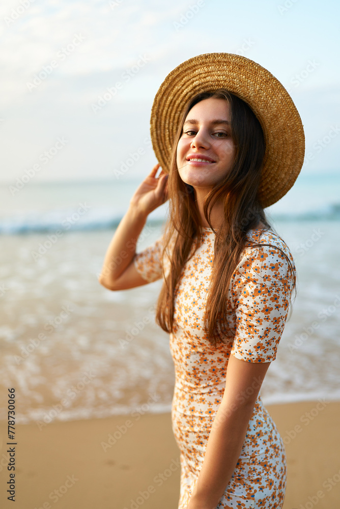 Smiling young woman in floral dress and straw hat stands on sandy beach. Fashionable female enjoys summer by sea at sunset. Vintage vibe, travel, leisure concept. Solo traveler poses at ocean edge.