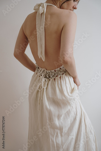 Back view of young woman in neutral cream beige evening dress against white wall. Minimal chic fashion concept