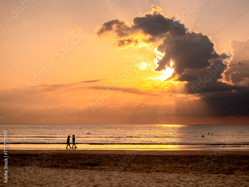 Fantastic sunset sky with beautiful sun rays behind the cloud over the sea water background with silhouette people on shore. Summer sand beach in evening time. Early morning sunrise or dusk seascape.
