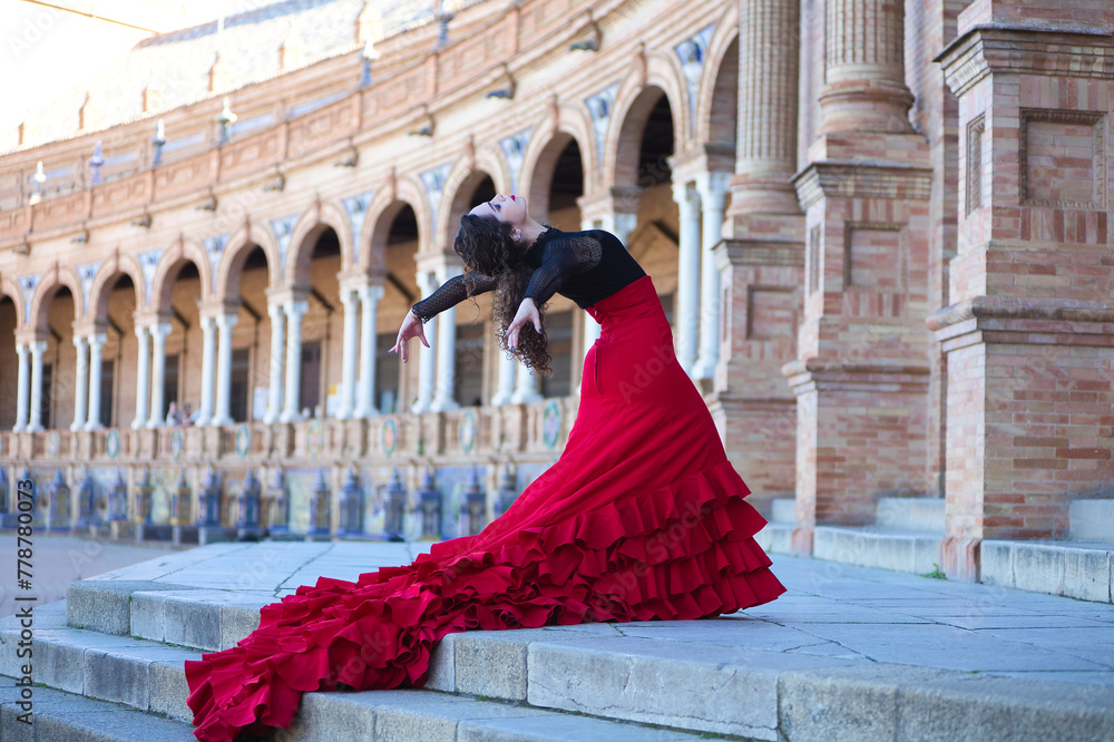 Beautiful woman dancing flamenco in a square in Seville, Spain. She is wearing a typical red and black gypsy dress and dancing flamenco with a lot of art. The dress hangs down the steps of the square.