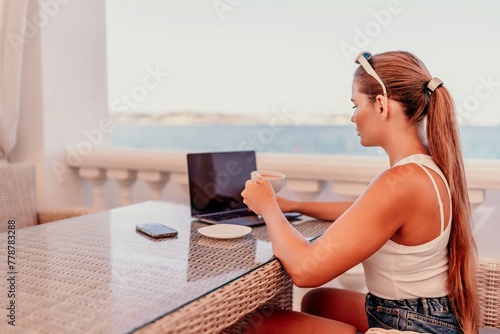 woman is sitting at a table with a laptop and a cup of coffee. She is wearing sunglasses and a ponytail. The scene suggests a relaxed and leisurely atmosphere
