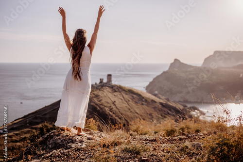 A woman in a white dress stands on a rocky hill overlooking the ocean. She is smiling and she is happy.