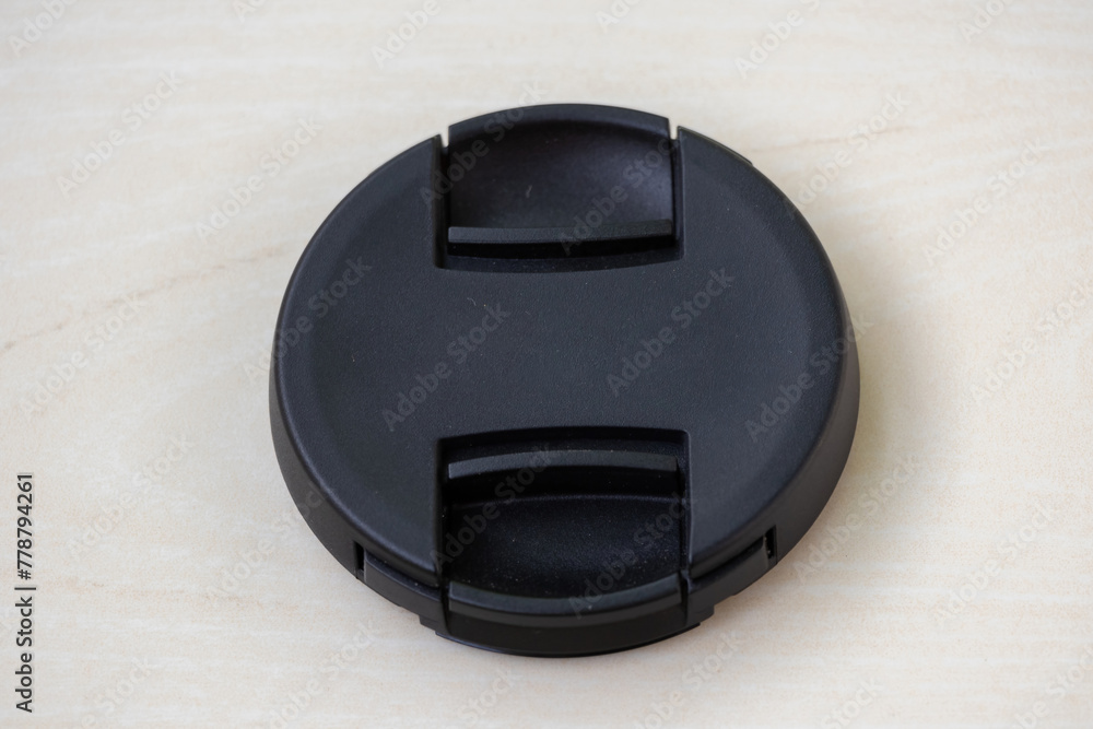 Close-up view of a black camera lens cap without logo on wooden background. A lens cover or lens cap protects camera lenses from scratches, dust, rain, snow, minor collisions, and oily residue. Close-up view of a black camera lens cap without logo on wooden background. A lens cover or lens cap protects camera lenses from scratches, dust, rain, snow, minor collisions, and oily residue.