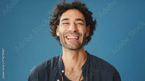 Close up, laughing attractive man with curly hair, dressed in blue shirt, crossing his arms and looking at camera isolated on blue background in studio
