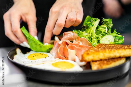 A chef garnishes a gourmet breakfast plate with fresh green salad, showcasing vibrant colors and textures of eggs, meat, and vegetables