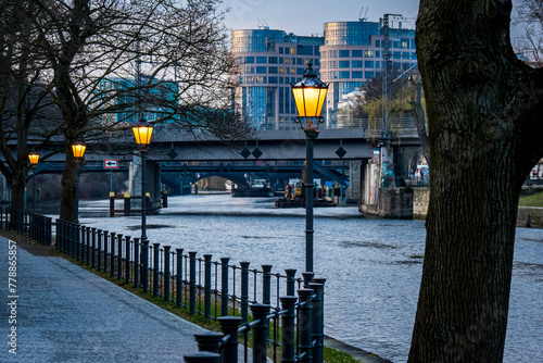 In February evenings Bellevue Ufer waterfront offers a serene view of Spree river, adorned with glowing street lamps, leading to sunlit office buildings in Berlin Moabit, flanked by the Railway bridge