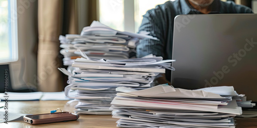 At the desk working person on laptop computer with a pile of documents on table in office. Overwork and excess responsibilities at work