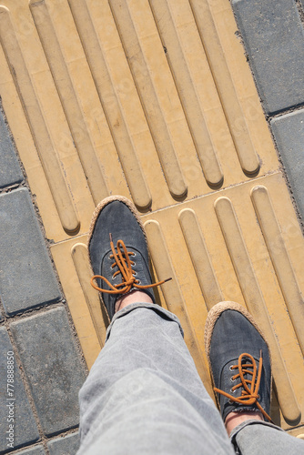 Women's feet in sneakers stand on a tactile tile. The road for the blind.	