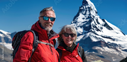 A photo of a middle-aged man and woman in a red jacket, black sunglasses with backpacks smiling at the camera standing on Zermatt mountain with Matterhorn behind them
