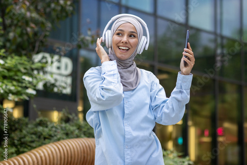 A cheerful woman in a hijab listens to music with headphones and dances joyfully outside an office building. © Liubomir
