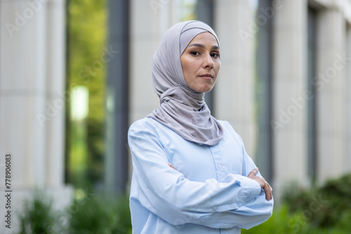 A professional businesswoman in hijab stands confidently outdoors near modern office buildings.