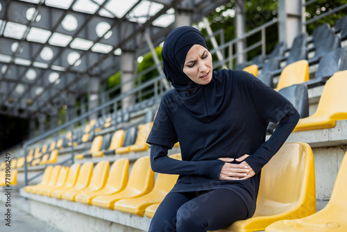 A female athlete wearing a hijab experiences discomfort and stomach pain while sitting alone in a stadium with empty seats. photo