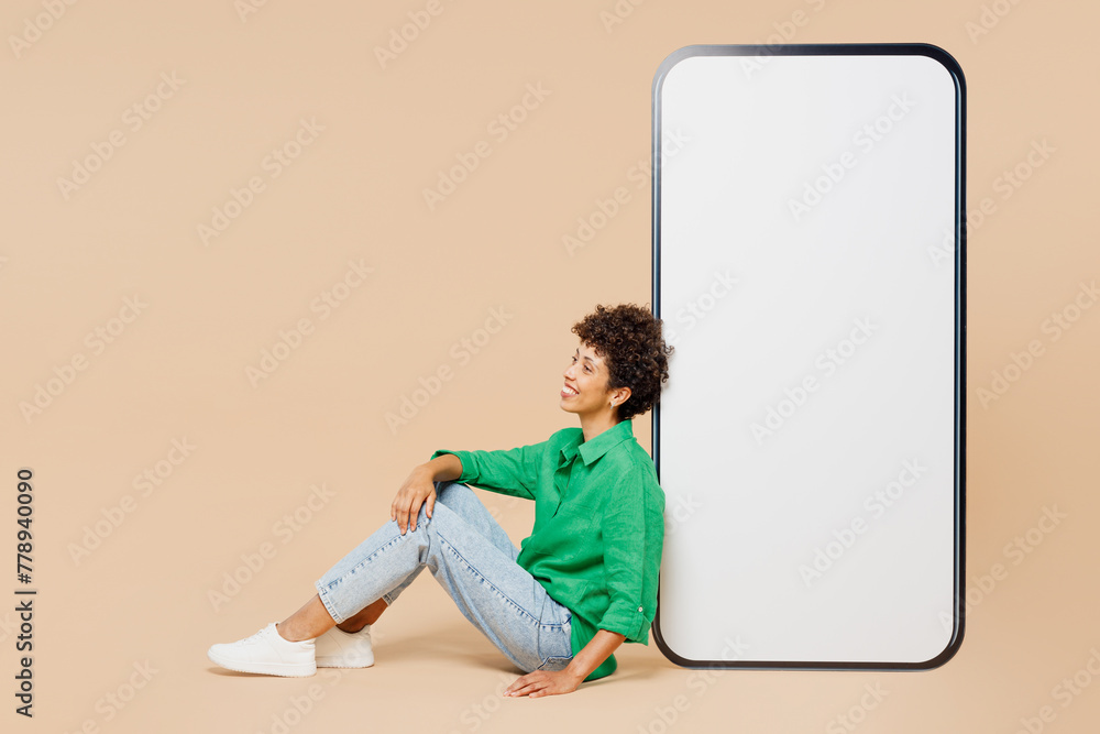 Full body side view young woman of African American ethnicity wear green shirt casual clothes sits near big huge blank screen mobile cell phone smartphone with area isolated on plain beige background.