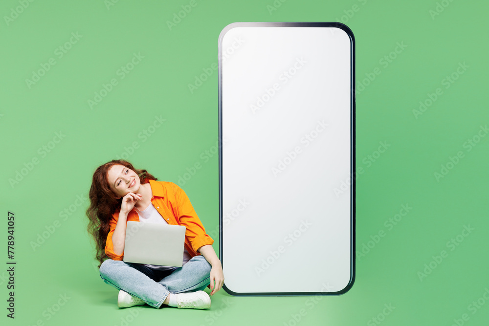 © ViDi Studio - Full body young IT woman wear orange shirt white t-shirt casual clothes big huge blank screen mobile cell phone smartphone use laptop pc computer isolated on plain green background. Lifestyle concept.