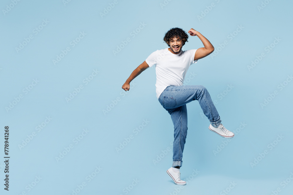 Full body young happy Indian man he wear white t-shirt casual clothes doing winner gesture celebrate clench fists raise up leg isolated on plain pastel light blue cyan background. Lifestyle concept Full body young happy Indian man he wear white t-shirt casual clothes doing winner gesture celebrate clench fists raise up leg isolated on plain pastel light blue cyan background. Lifestyle concept