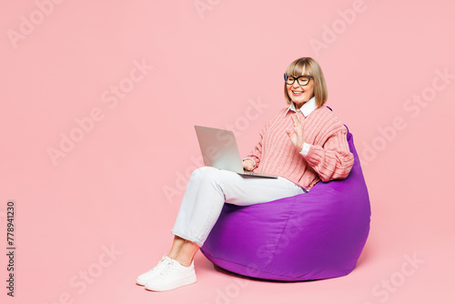 Full body elderly IT woman 50s years old wear sweater shirt casual clothes glasses sit in bag chair hold use work on laptop pc computer waving hand isolated on plain pink background Lifestyle concept