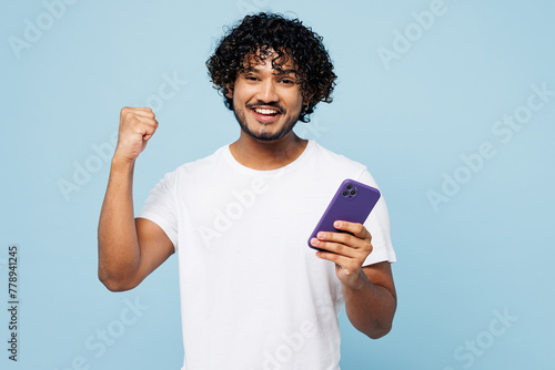 Young happy Indian man he wear white t-shirt casual clothes hold in hand use mobile cell phone do winner gesture isolated on plain pastel light blue cyan background studio portrait. Lifestyle concept.