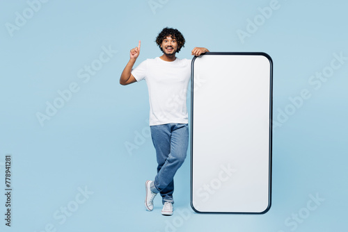 Full body young Indian man wear white t-shirt casual clothes big huge blank screen mobile cell phone smartphone with area point finger up isolated on plain pastel blue background. Lifestyle concept.