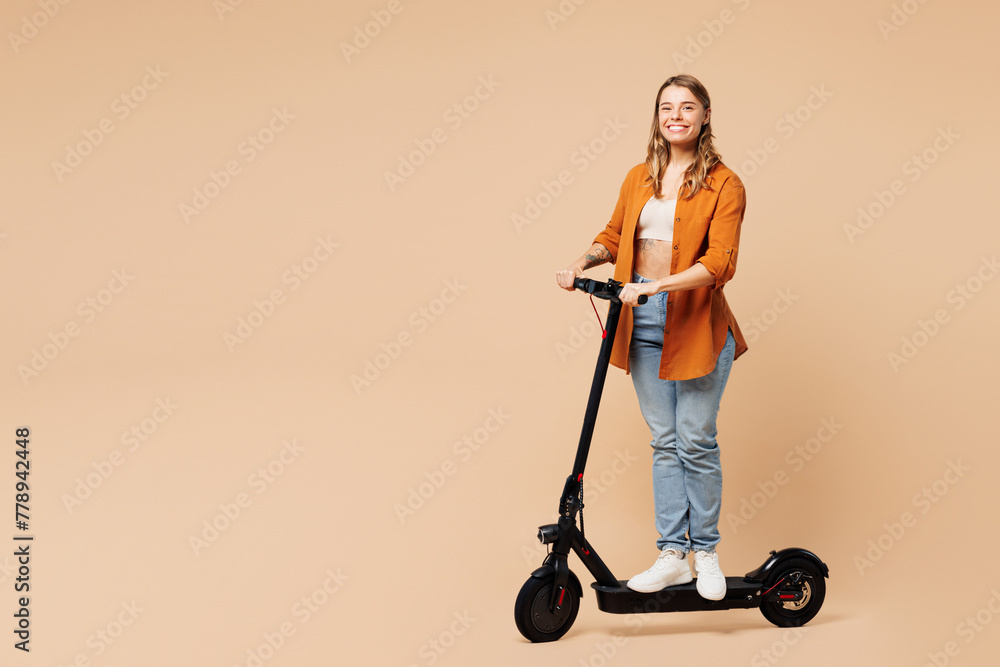 Full body side view smiling happy young woman she wear orange shirt casual clothes ride electric scooter look camera isolated on plain pastel light beige background studio portrait. Lifestyle concept. Full body side view smiling happy young woman she wear orange shirt casual clothes ride electric scooter look camera isolated on plain pastel light beige background studio portrait. Lifestyle concept.