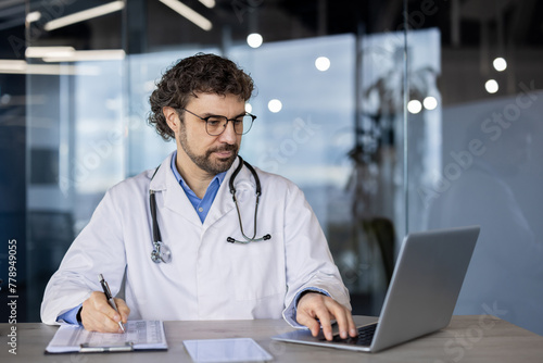 A professional male doctor in a lab coat using a laptop and writing notes on a clipboard in a modern healthcare setting.