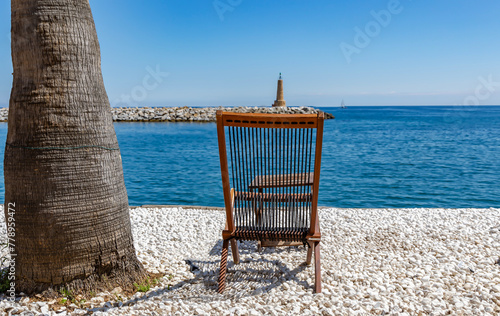 Wooden sun lounger on the Mediterranean shore near a palm tree overlooking the lighthouse. Peaceful holiday concept.