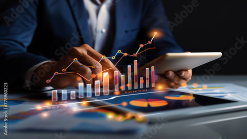 A businessman's hand using a tablet with a financial graph and bar chart of stock market growth on a desk with an office background. A concept for investment