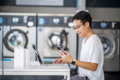 Young Asian man using laptop and smartphone doing his work while waiting for washing machine working at laundry station  Man with housework and laundry day.