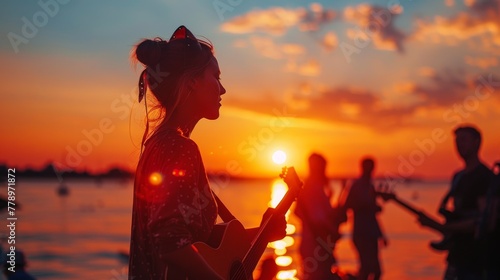 A woman is playing a guitar on a beach at sunset. There are other people in the background, and the scene has a relaxed and peaceful mood