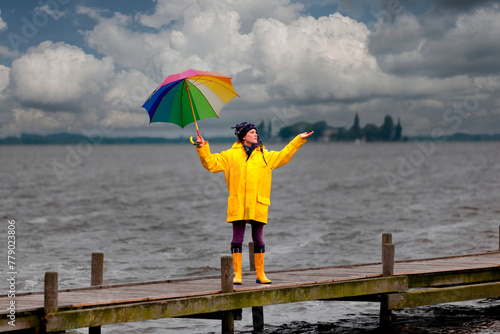 Junge Frau mit gelber Regenjacke und buntem Schirm auf einem Steg