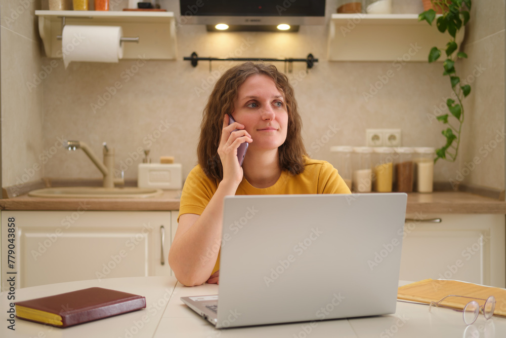 A woman with a laptop is talking on a mobile phone while sitting at a table in a home kitchen. An adult female businesswoman works from home, a remote office A woman with a laptop is talking on a mobile phone while sitting at a table in a home kitchen. An adult female businesswoman works from home, a remote office