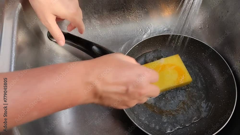 Unas manos de mujer lavando  un sartén  con una esponja amarilla con jabón líquido y  agua en un fregadero