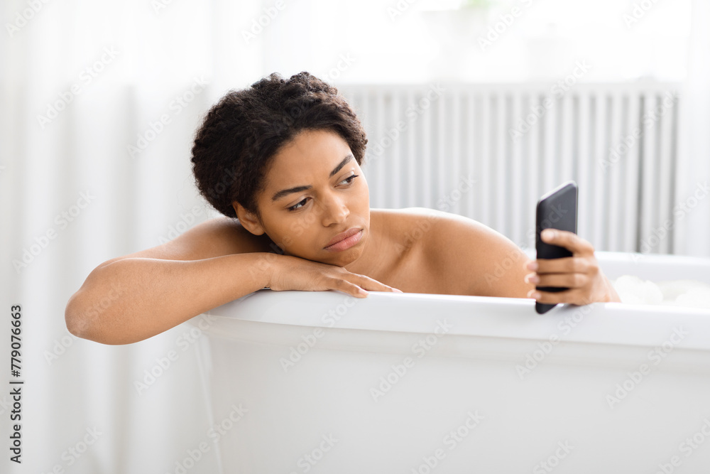 Pensive African American woman in bathtub with phone Pensive African American woman in bathtub with phone