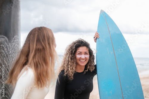 A banner with two young female surfers talking next to their surfboards. Portrait of a Latino woman with curly hair who smiles, her face has colorful zinc and she holds a blue surfboard with her hand.