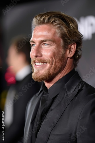 A smiling handsome man with light facial hair and goatee, blue eyes, in a black suite at a red carpet event looking to the side.