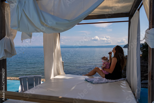 Woman holding a baby in lounger canopy beach bed in luxury hotel with scenic view of idyllic coastline of Gulf of Piran, Adriatic Mediterranean Sea, Slovenia, Europe. Summer seaside family vacation photo