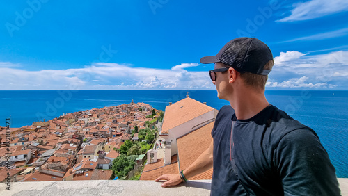 Tourist man enjoying scenic aerial view of coastal town Piran, Primorska, Slovenia. Shimmering azure waters of Adriatic Sea. Tranquil Mediterranean atmosphere. Exploring Slovenian Coast in Istria photo
