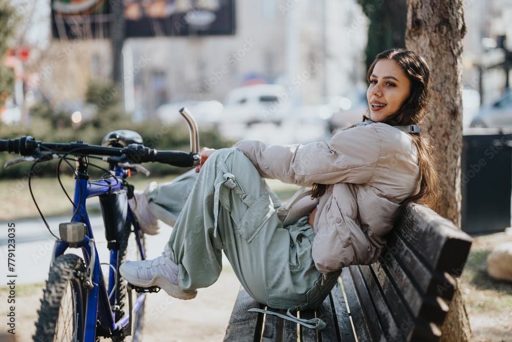 A smiling young female sits on a bench in the park, taking a leisurely break beside her mountain bike, radiating casual urban lifestyle vibes.