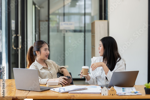 Two smiling female colleagues work together on a project with a laptop and coffee in office space.