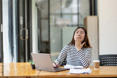 A young woman yawns while using a laptop at her desk. being tired during a busy work day © Wasana