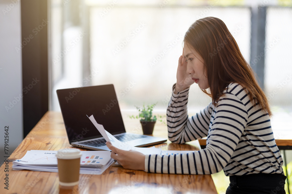 A worried businesswoman looking at laptop screen with documents spread out, concern over work problems. A worried businesswoman looking at laptop screen with documents spread out, concern over work problems.