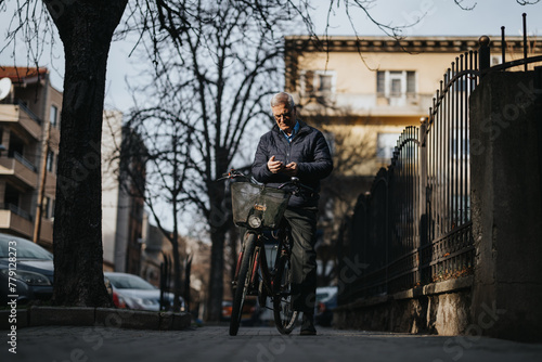 Mature man checks phone messages in quiet street, with bike. Image evokes urban lifestyle, active seniors and sustainable transportation.