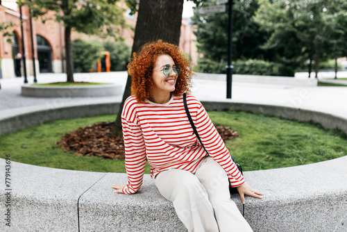 Happy cheerful millennial curly-haired redhead female sitting on stoned bench in city park, waiting for her friend to come after long time not seeing each other, enjoying fresh air and beautiful view