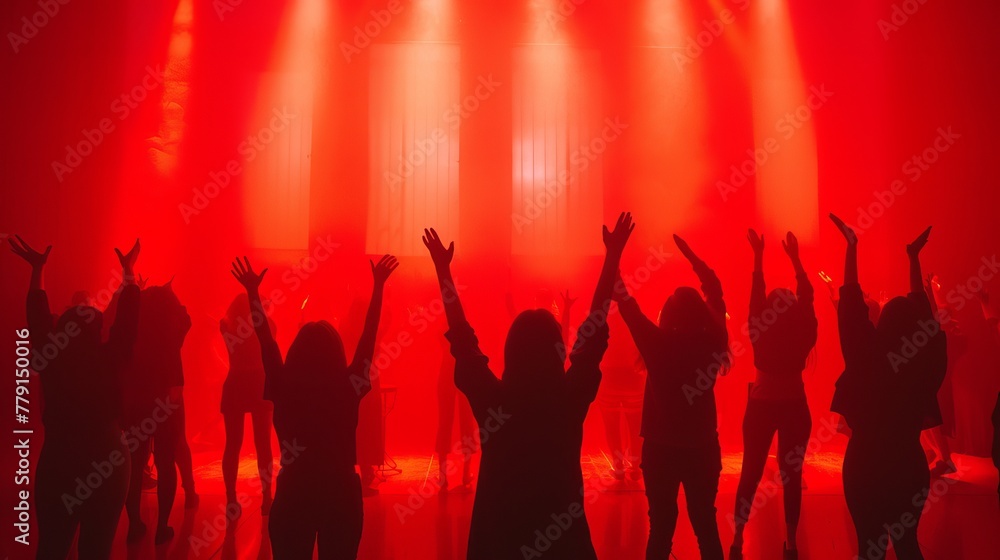 People stand in silhouette with arms raised in a concert hall bathed in red light.